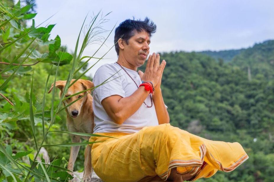 Lead Yoga Teacher Anoop meditating outdoors with folded hands in a peaceful green mountain setting.