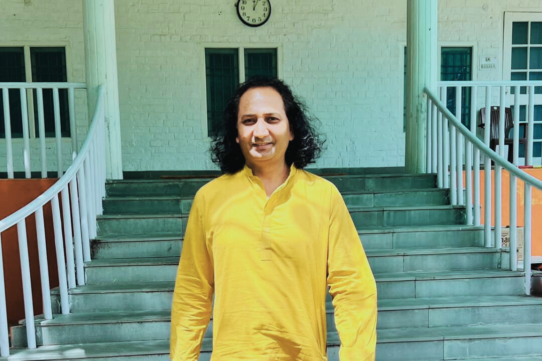 A man in a yellow traditional kurta standing in front of a staircase, smiling calmly.