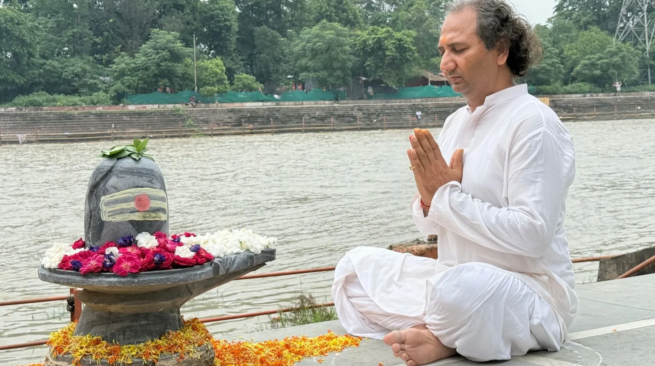 Man practicing meditation and pranam mudra beside a Shiva Lingam at the riverside.